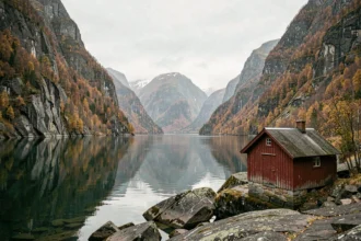 Paysage de fjord norvégien en automne avec cabane en bois rouge et eaux calmes reflétant les falaises rocheuses