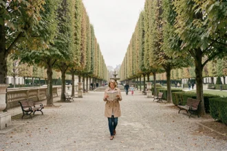 Allée arborée d'un parc parisien classique avec une femme se promenant sur un chemin de gravier entouré de marronniers taillés et de bancs en fer forgé